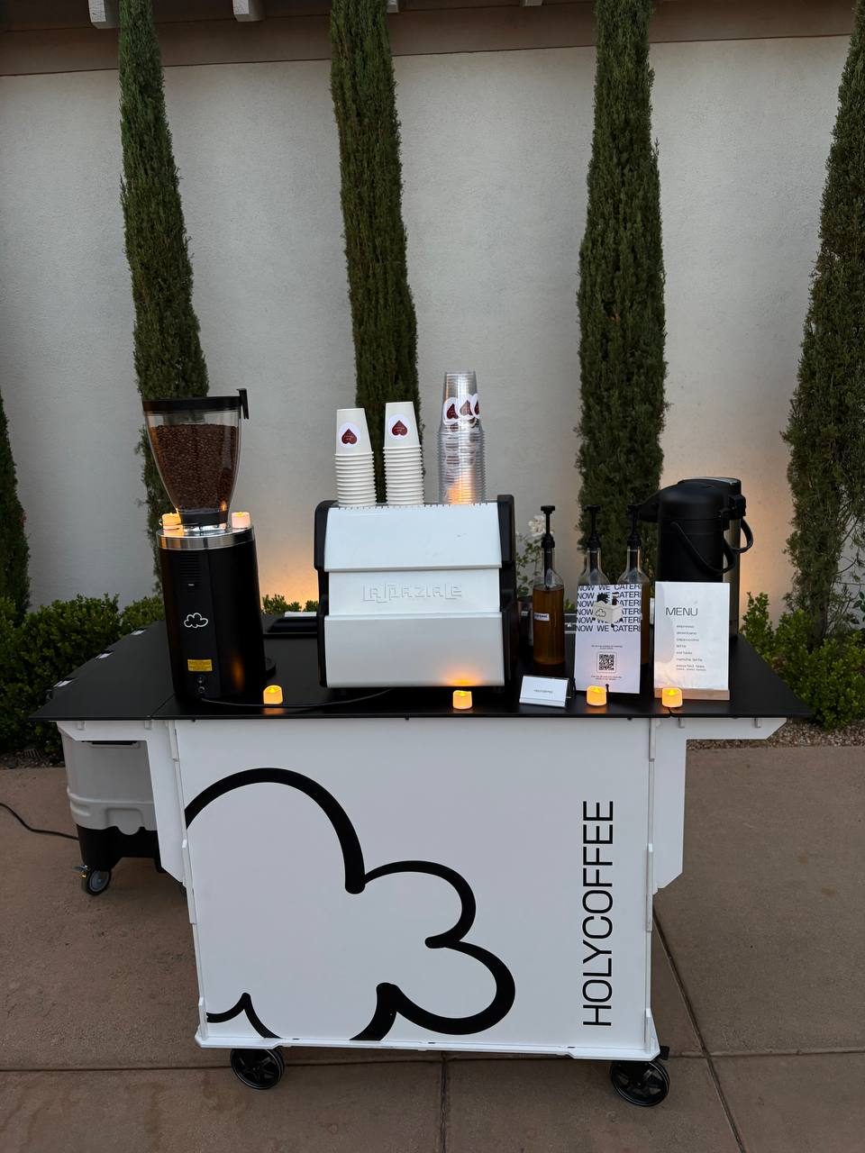 Holy Coffee cart with candles in front of tall cypress trees