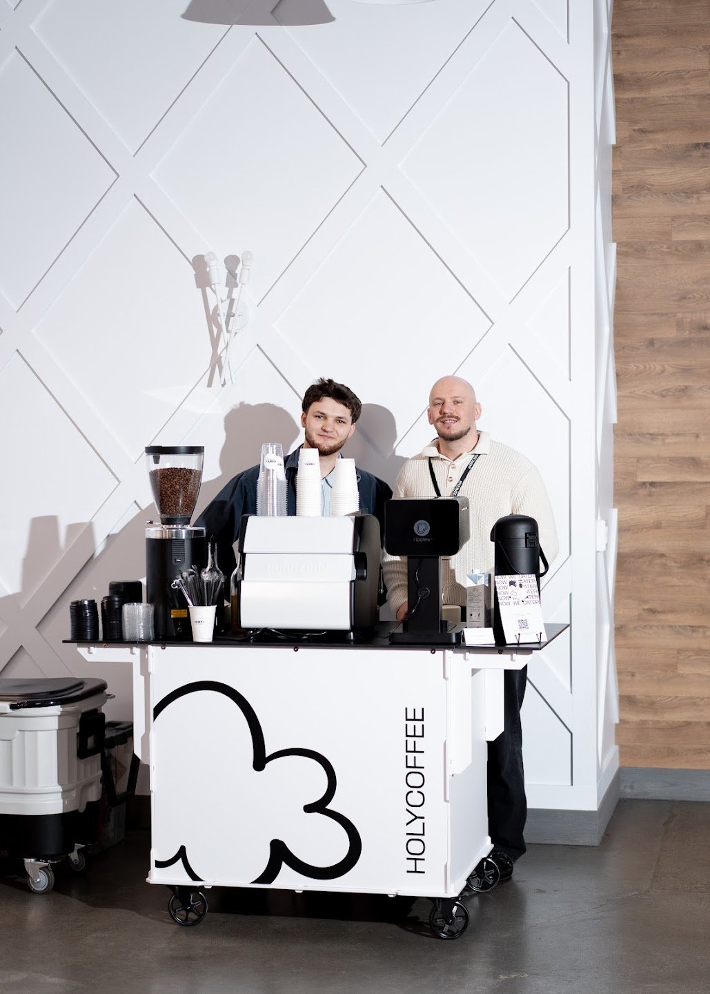 Two baristas at the Holy Coffee cart against a white paneled wall