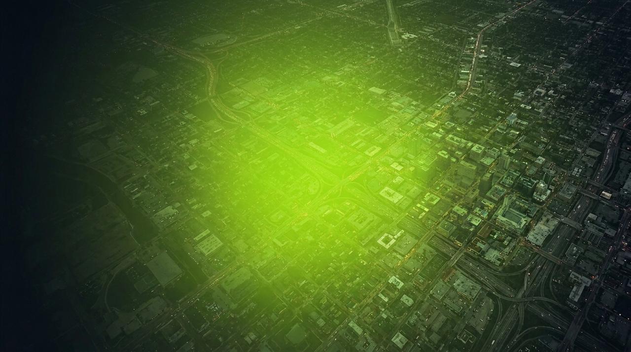 Aerial night view of the same city with bright green light now spreading across almost the entire frame, reaching the far edges