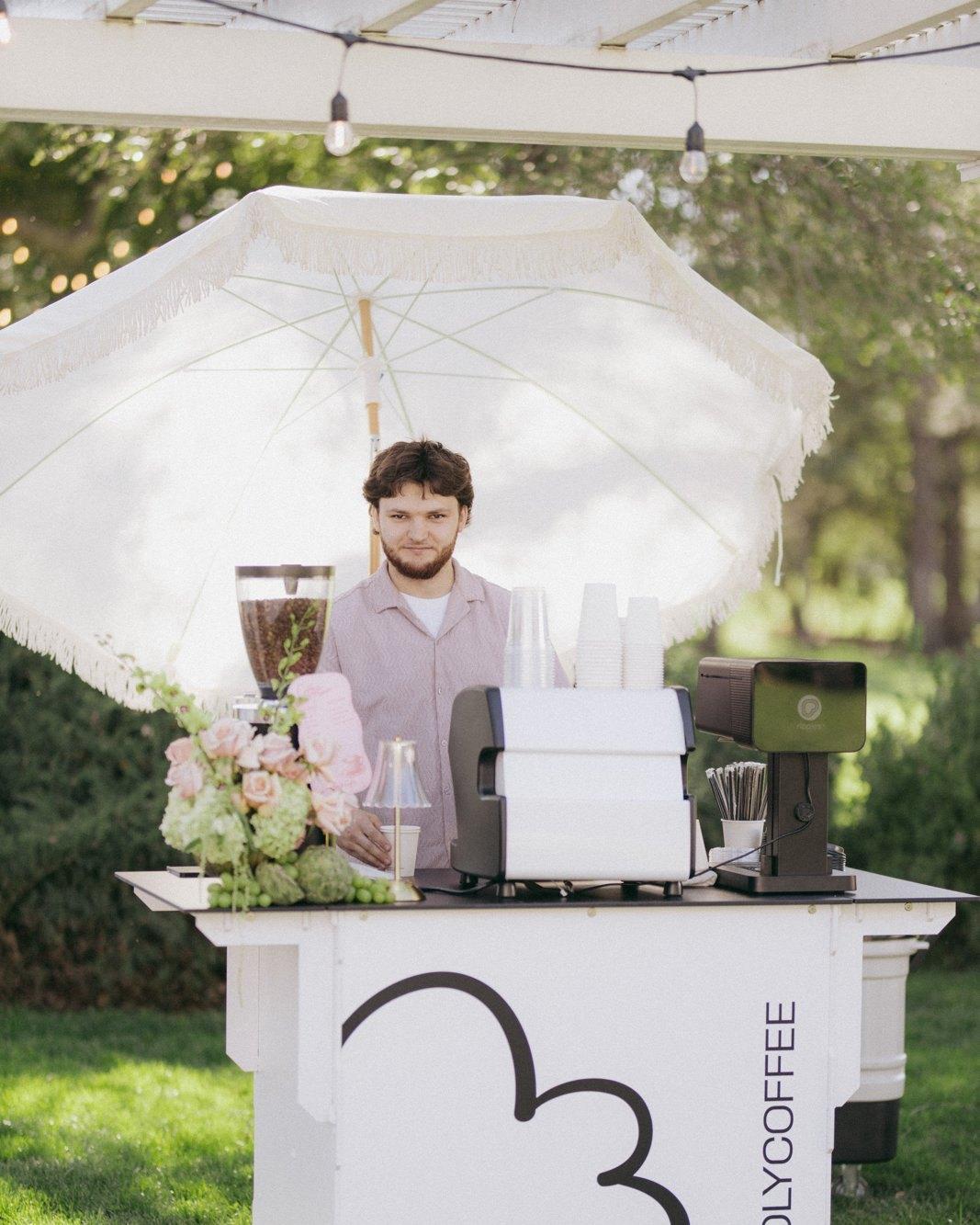 A Holy Coffee Cart barista at the cart, under a white umbrella with flowers, on a lawn