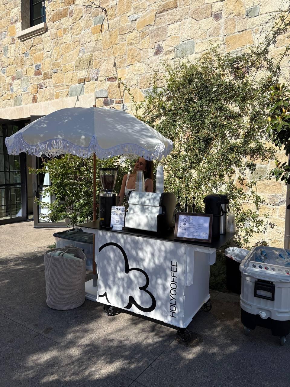 Holy Coffee cart with a fringed umbrella by a stone venue wall