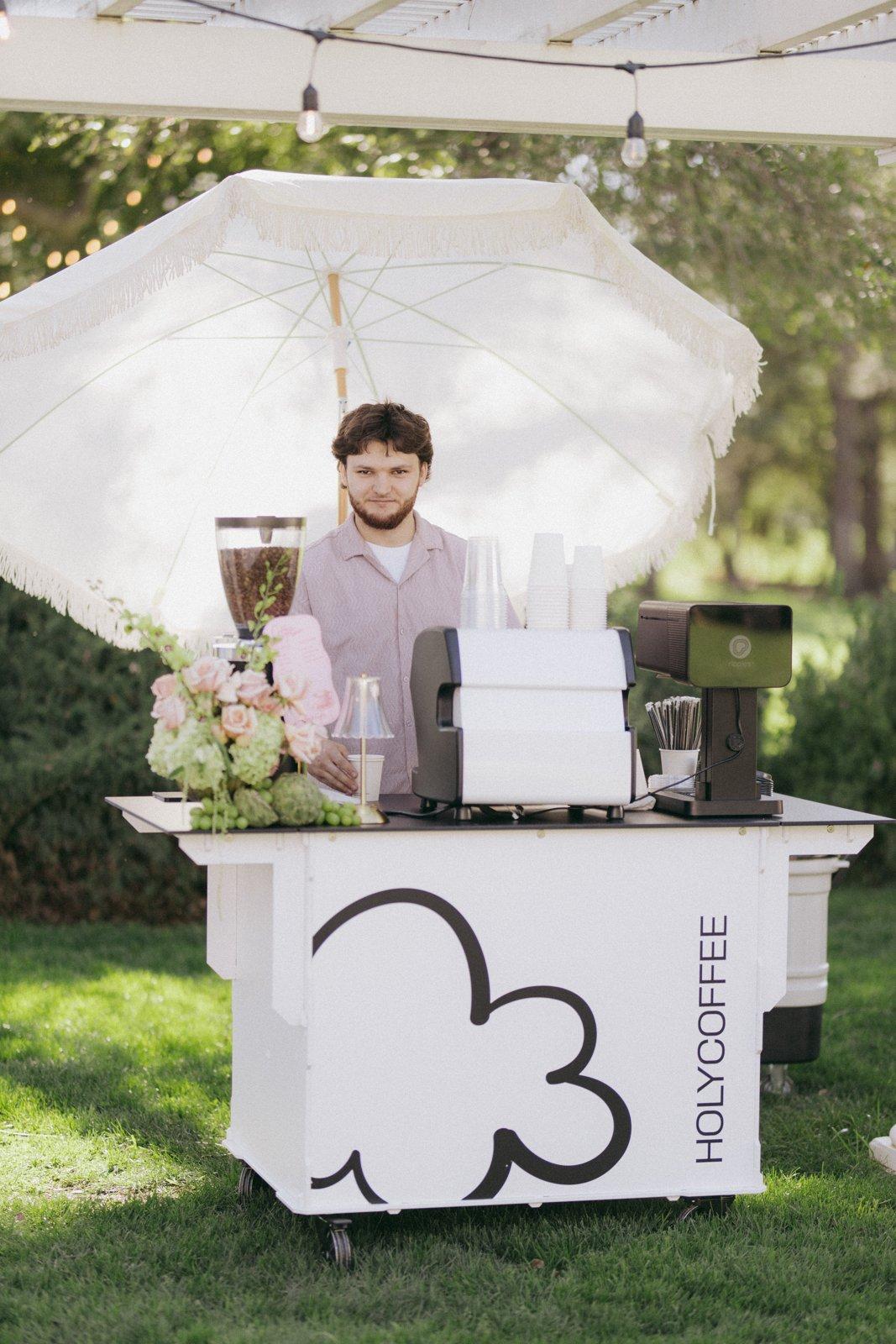 Holy Coffee cart with a white umbrella and flowers on a wedding lawn
