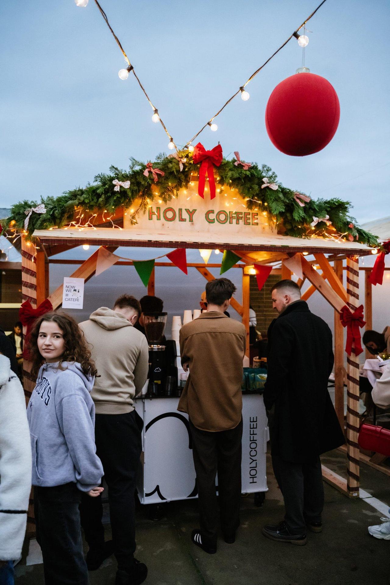 The HOLYCOFFEE cart under a holiday booth with garland, red bows and string lights while guests line up for coffee
