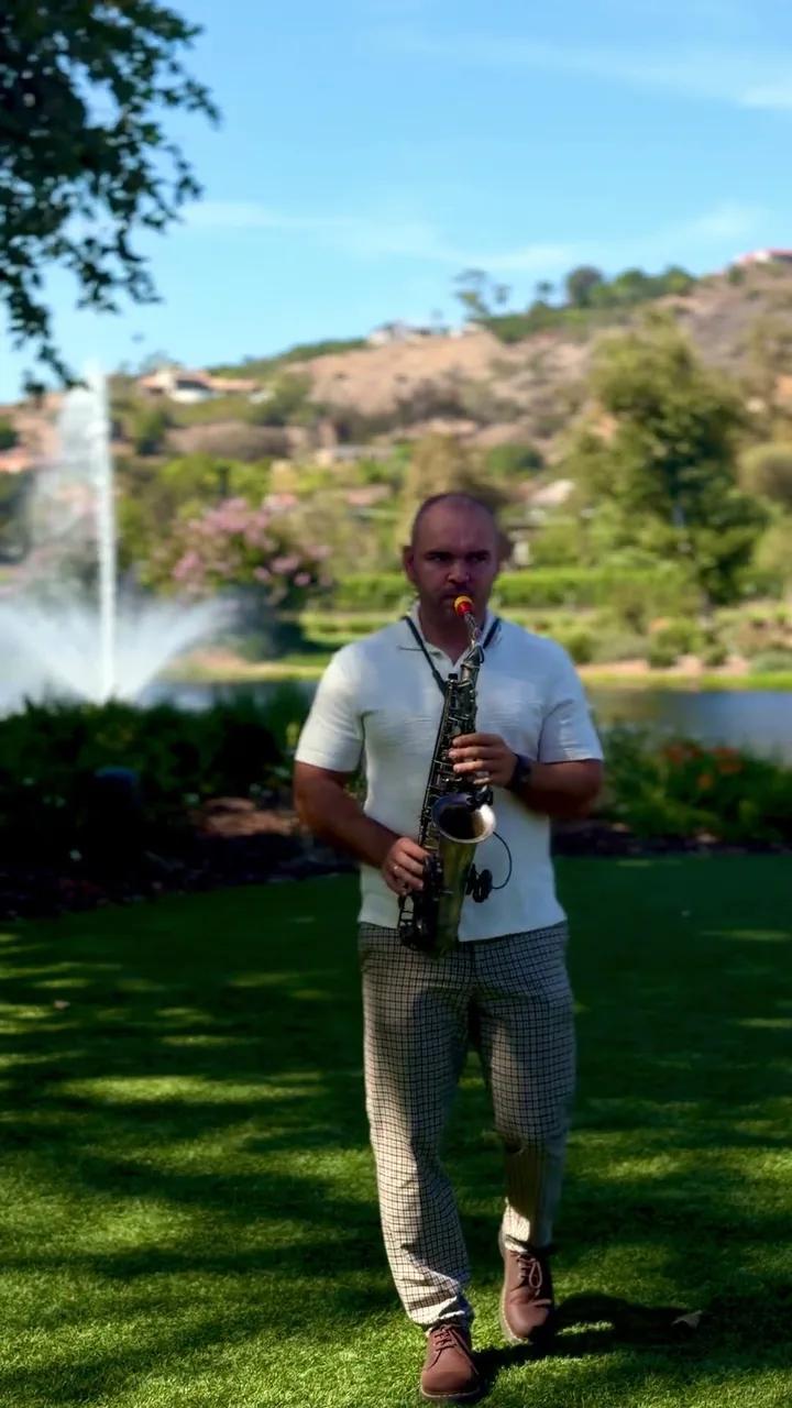 Igor playing sax on a lawn in front of a fountain and green hills on a sunny day