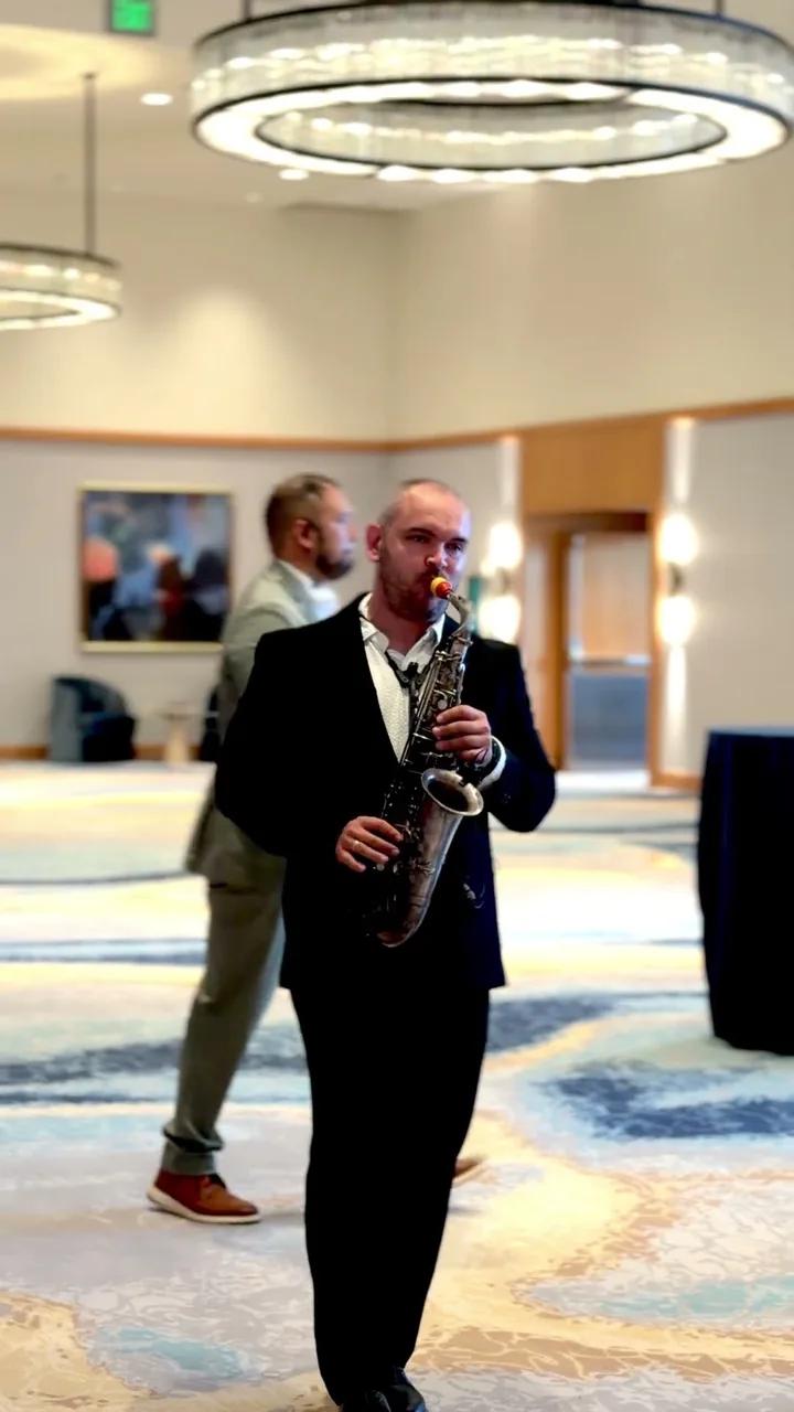Igor in a black suit playing sax in a ballroom under a round chandelier