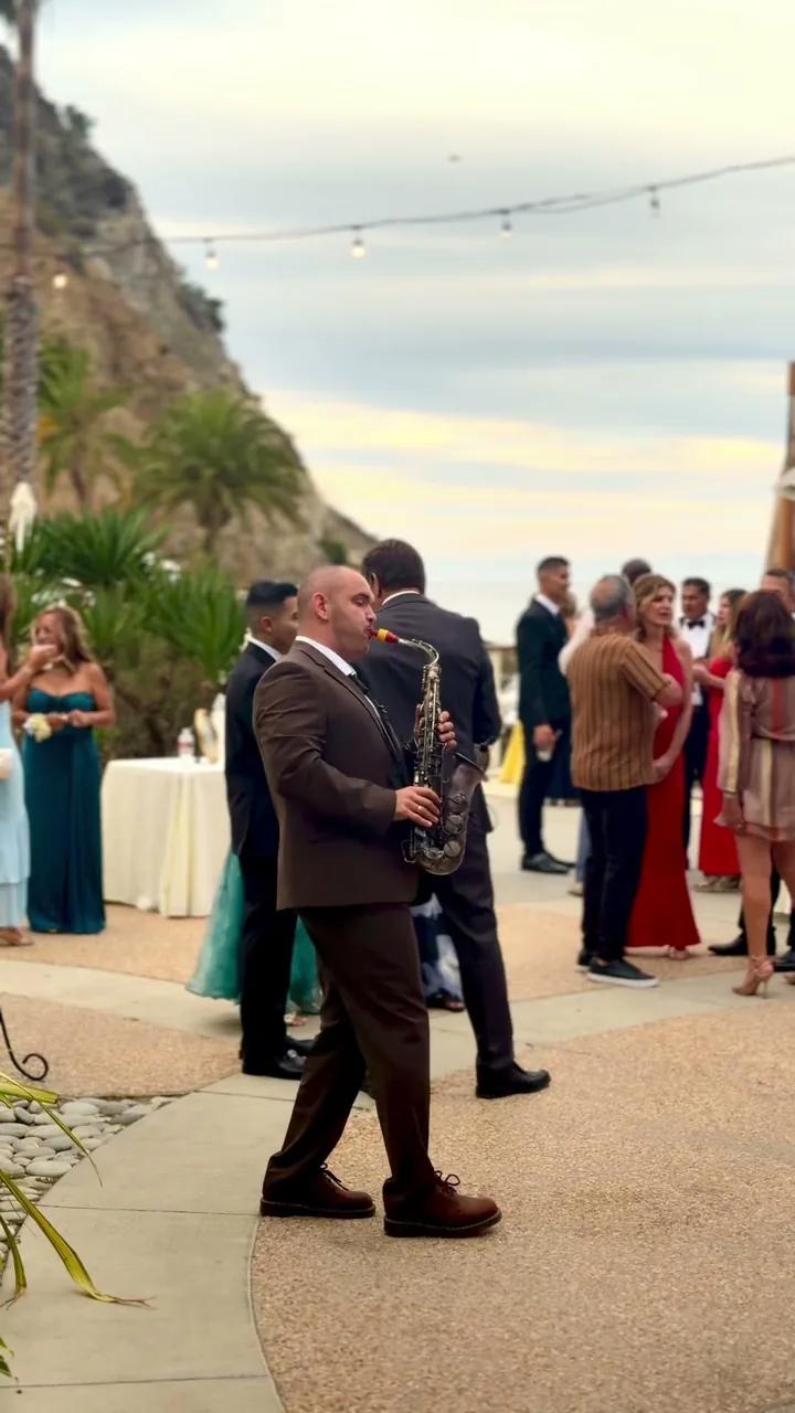 Igor in a brown suit playing sax as guests dance on a seaside terrace with string lights