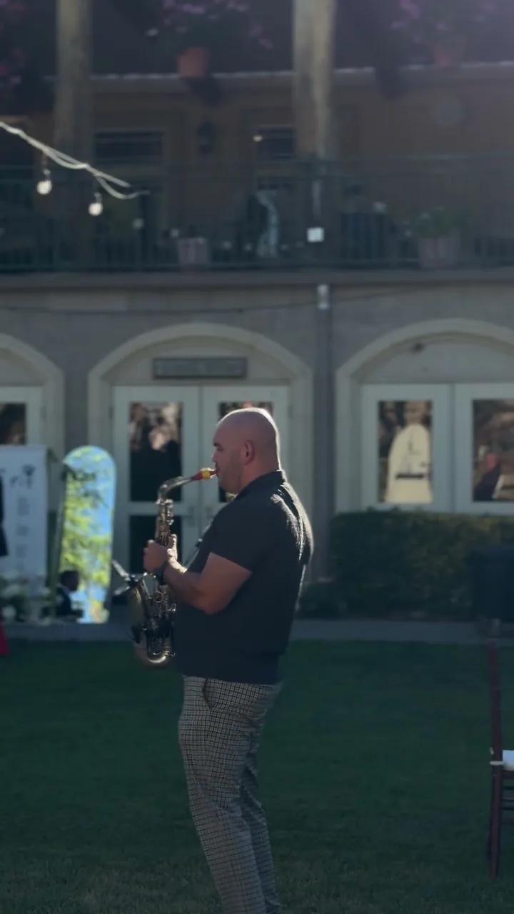 Igor playing sax at dusk on a lawn among wedding guests in front of an arched venue