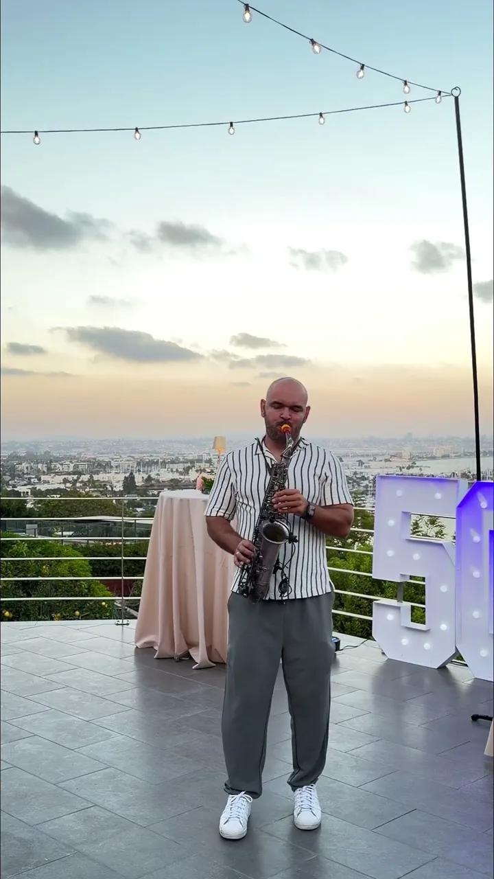 Igor playing sax on a rooftop at sunset at a private party, city lights and a balloon number behind
