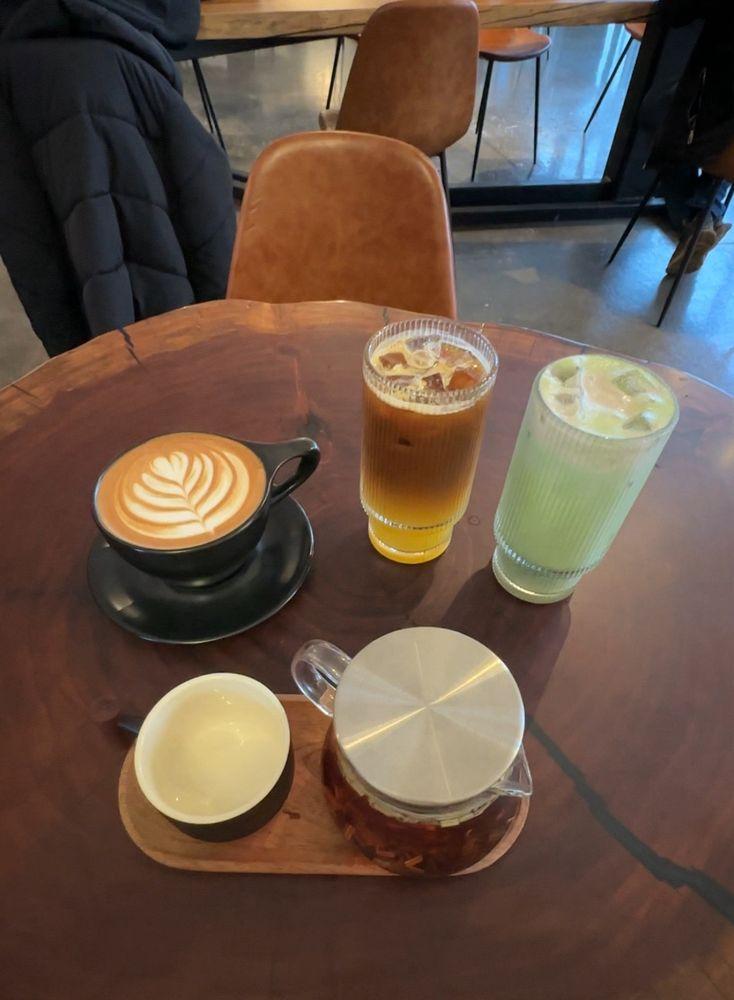 A latte, an espresso tonic, an iced matcha and a pot of tea on a wood table, leather chairs behind